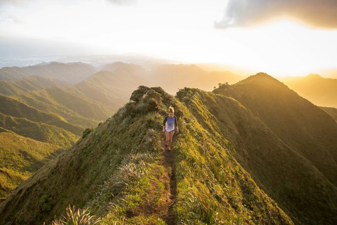 Persona caminando por una cresta montañosa bajo un cielo dorado al atardecer.