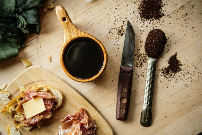 Tostada con mantequilla, jarra de salsa, cuchillo y café molido sobre una mesa de madera.