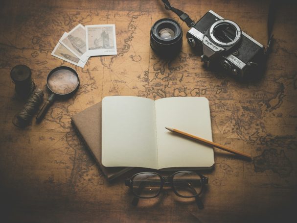 Mapa antiguo, cámara, gafas, lupa y cuaderno con lápiz sobre una mesa de madera.